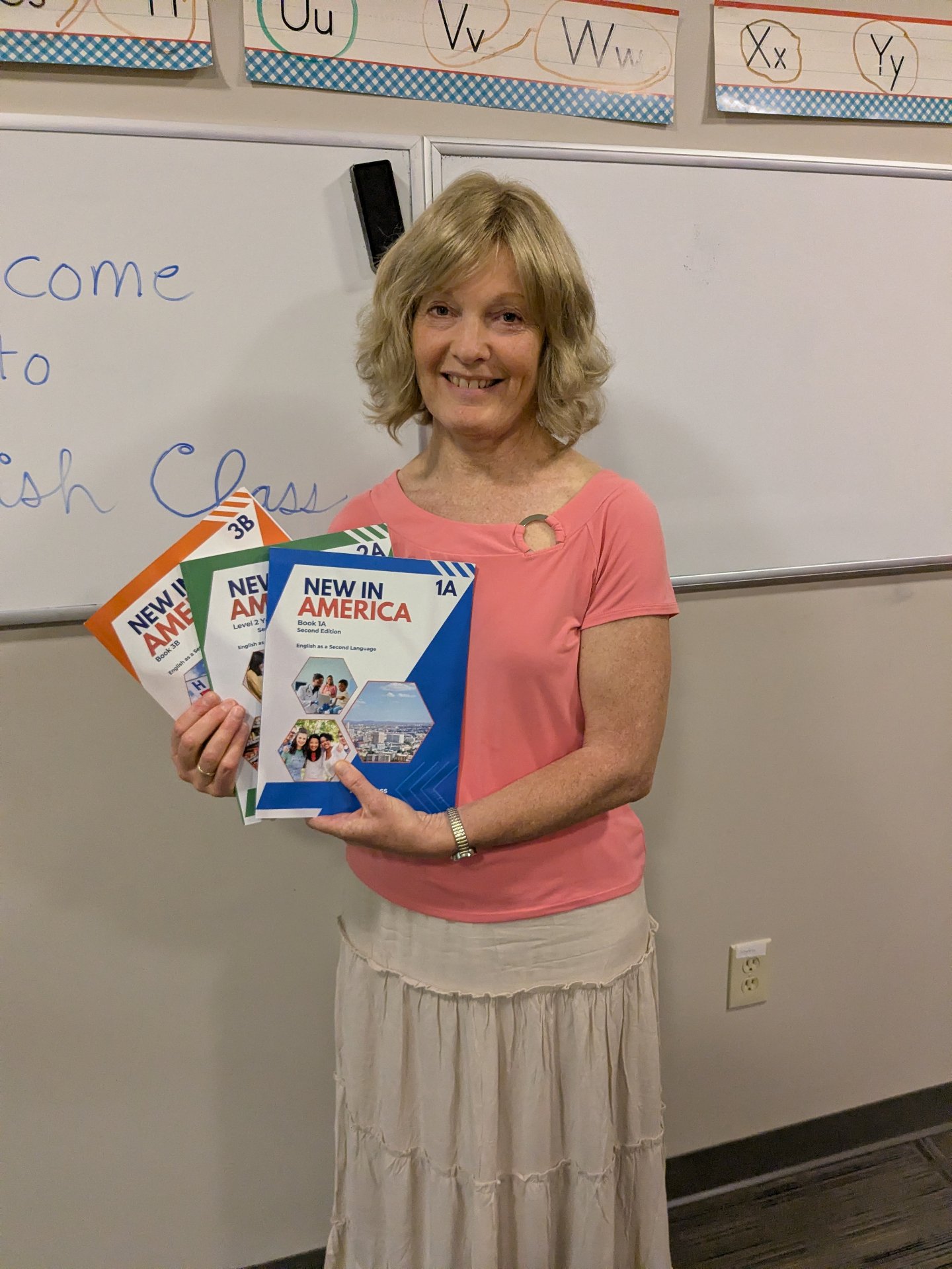An ESL teacher smiling in her classroom holding the New in America Book 1A, 2A, and 3B workbooks in front of a Welcome to English Class whiteboard